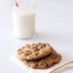 Two perfect chocolate chip cookies on a napkin with a glass of milk in the background.
