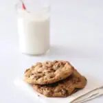 Two perfect chocolate chip cookies on a napkin with a glass of milk in the background.