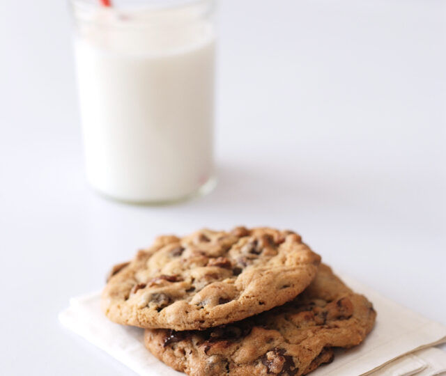 Two perfect chocolate chip cookies on a napkin with a glass of milk in the background.