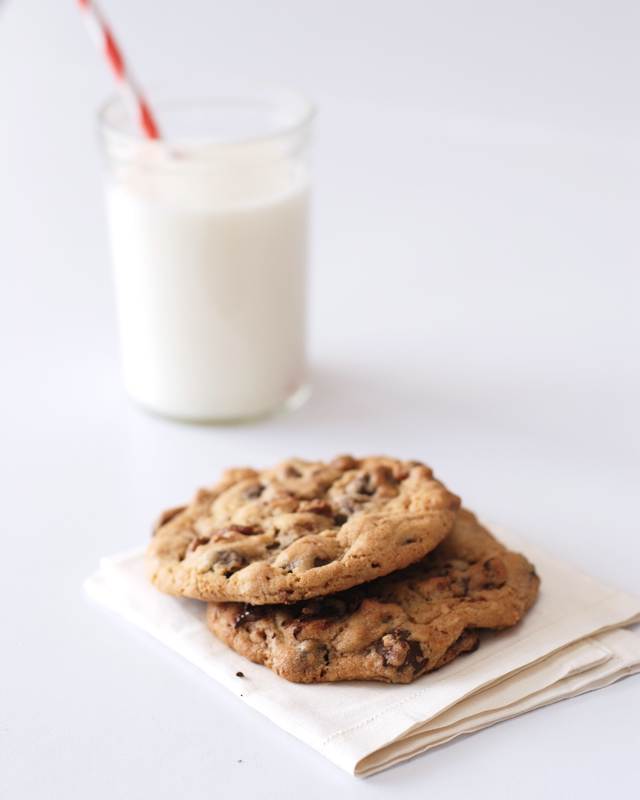 Two perfect chocolate chip cookies on a napkin with a glass of milk in the background.