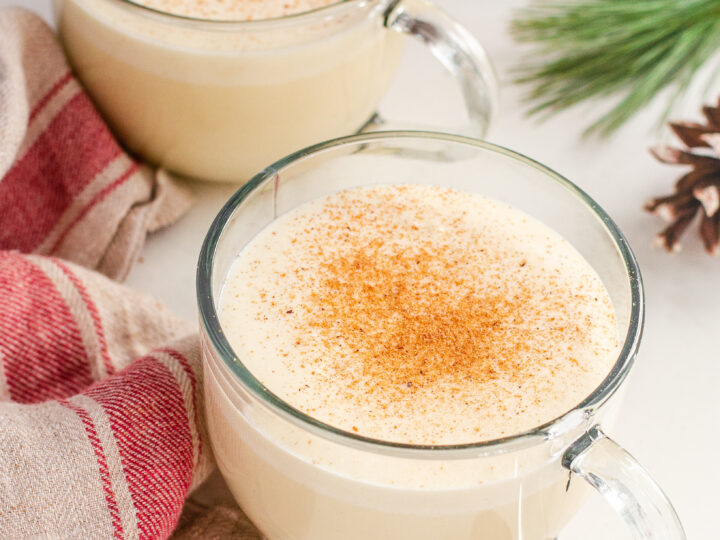 Two mugs of cooked eggnog with no alcohol with whole nutmegs, greenery, and a tea towel.