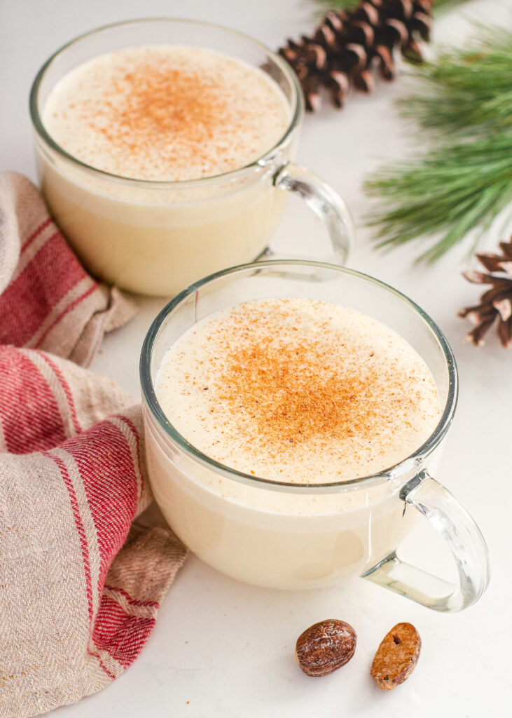 Tow mugs of my cooked eggnog recipe without alcohol with whole nutmegs, greenery, and a tea towel.