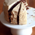 A chocolate layer cake with peanut butter frosting on a cake stand with a few pieces taken out.