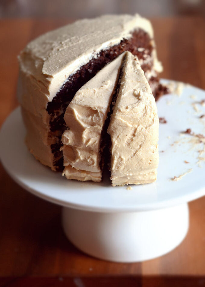 A chocolate layer cake with peanut butter frosting on a cake stand with a few pieces taken out.
