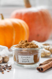 A jar of homemade pumpkin pie spice blend with pumpkins behind it and spiced around it.