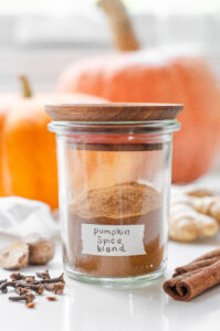 A jar of homemade pumpkin spice blend with pumpkin behind it and spiced around it.