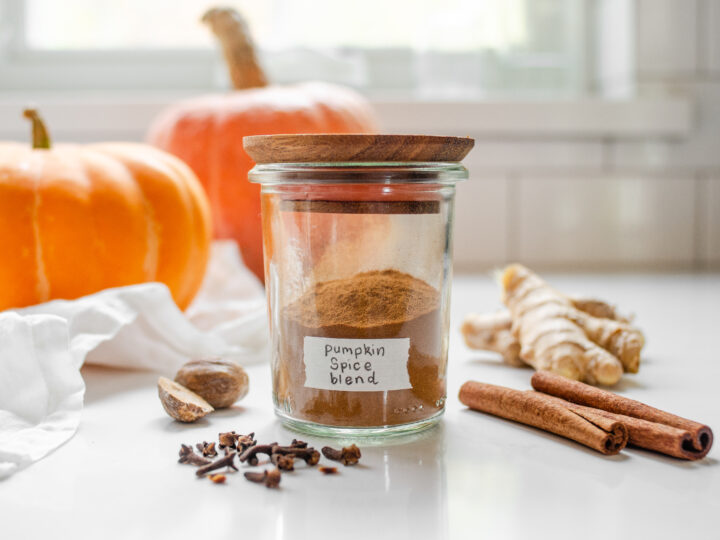 A jar of homemade pumpkin spice blend with pumpkin behind it and spiced around it.