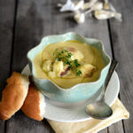 A bowl of roasted garlic cauliflower soup on a wooden background with slices of bread next to it.