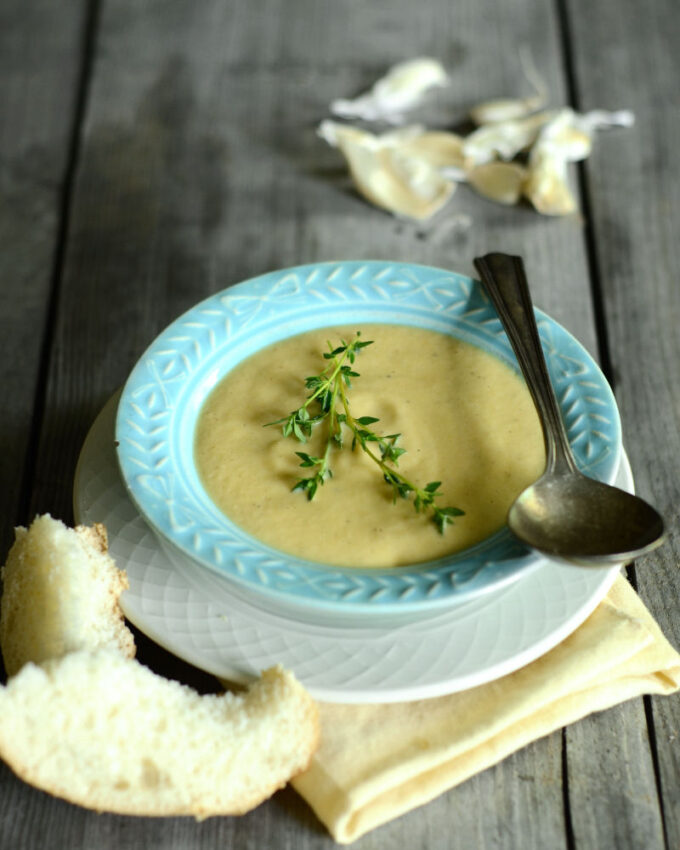 A bowl of roasted garlic cauliflower soup on a wooden background with slices of bread next to it.