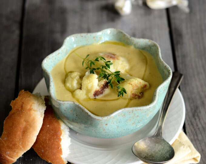 A bowl of roasted garlic cauliflower soup on a wooden background with slices of bread next to it.