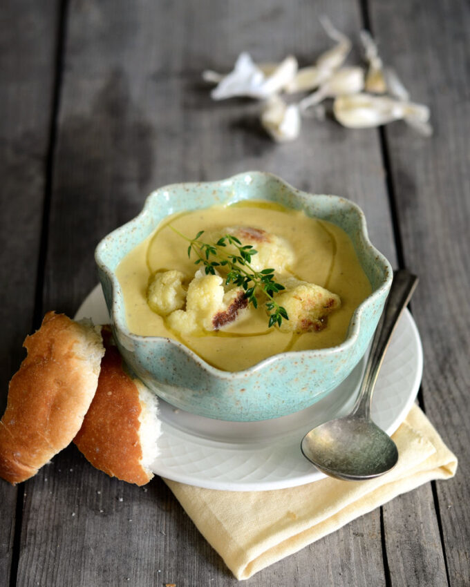 A bowl of roasted garlic cauliflower soup on a wooden background with slices of bread next to it.