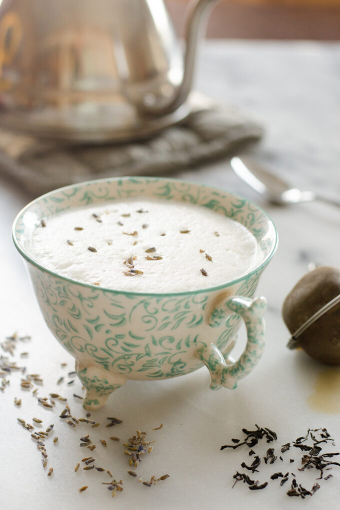 A teacup filled with a London Fog Tea Latte with a tea kettle in the background and a tea strainer off to the side.