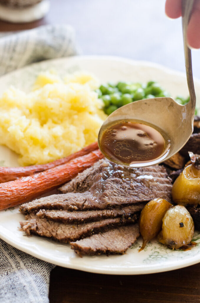 Pouring au jus over a plate of grass-fed roast beef chuck roast on a plate with mashed potatoes, green peas, and roasted veggies around it.