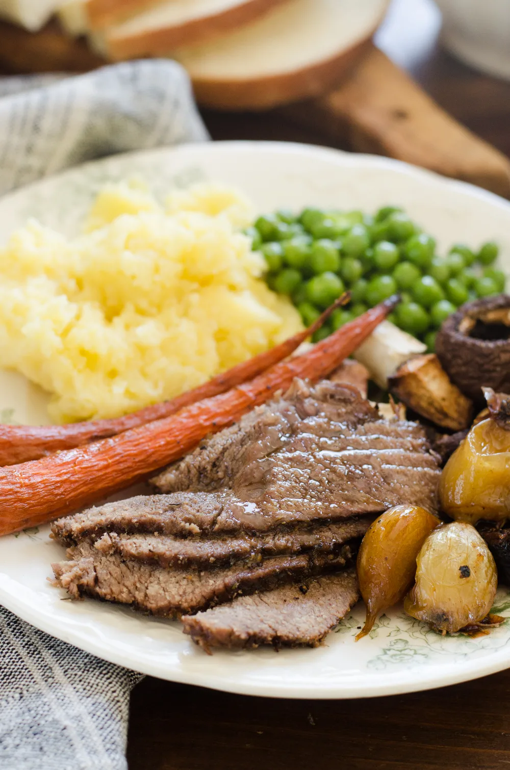 A plate of grass-fed roast beef chuck roast on a plate with mashed potatoes, green peas, and roasted veggies around it.