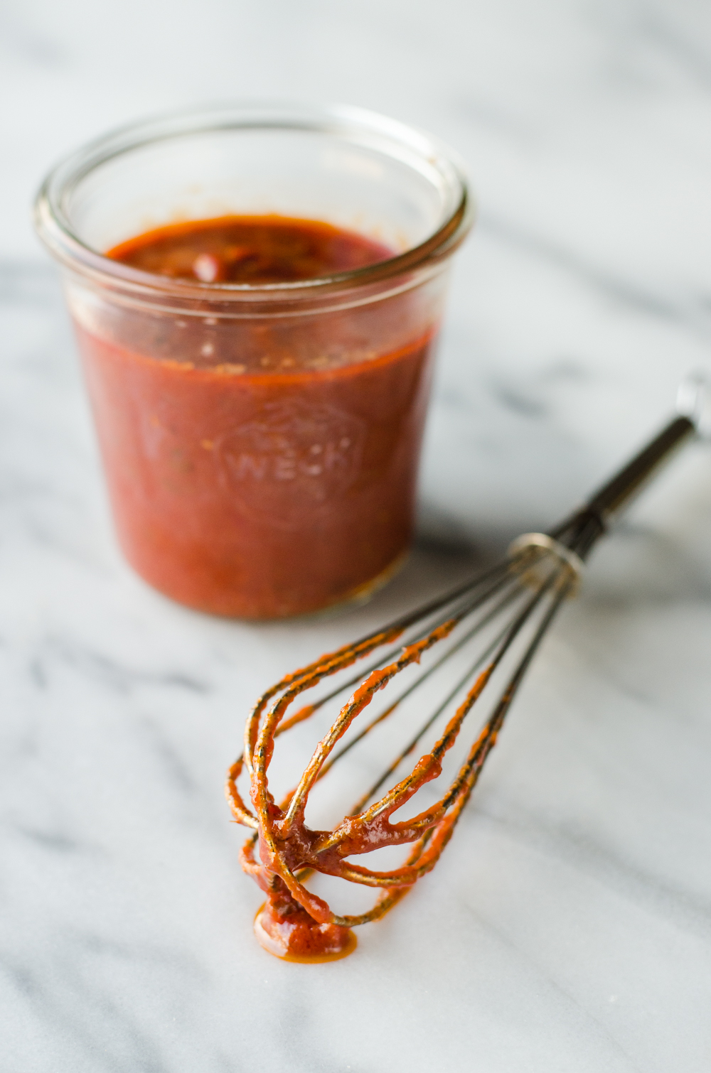 A jar of homemade pizza sauce made with tomato paste on a marble surface with a whisk in front of the jar.