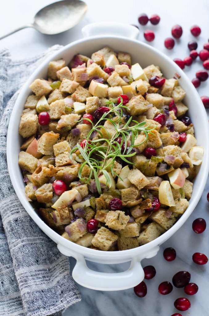 The baked Sourdough Stuffing in an oval casserole dish with a linen tea towel and cranberries off to the sides.