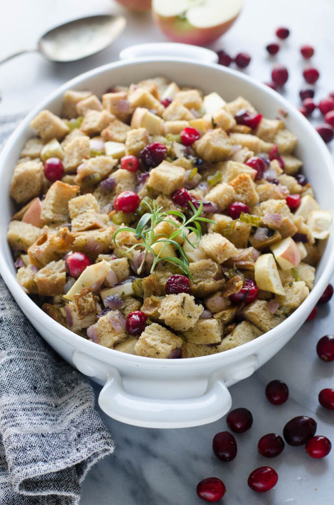 A casserole pan of Sourdough Stuffing made with cranberries and apples, with fresh rosemary on top, a linen tea towel off to the side, and fresh cranberries on the other side.