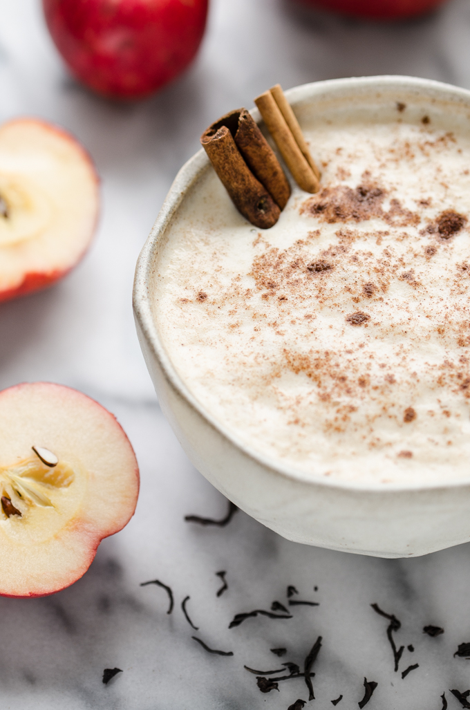 A closeup shot of a mug of Spiced Apple Tea Latte with cinnamon sticks inside and apple halves to the side.