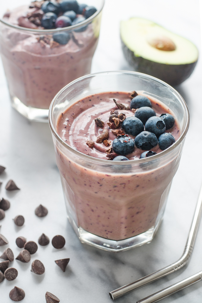 A chocolate blueberry avocado kefir smoothie on a marble surface with chocolate chips in the foreground and half an avocado in the background.