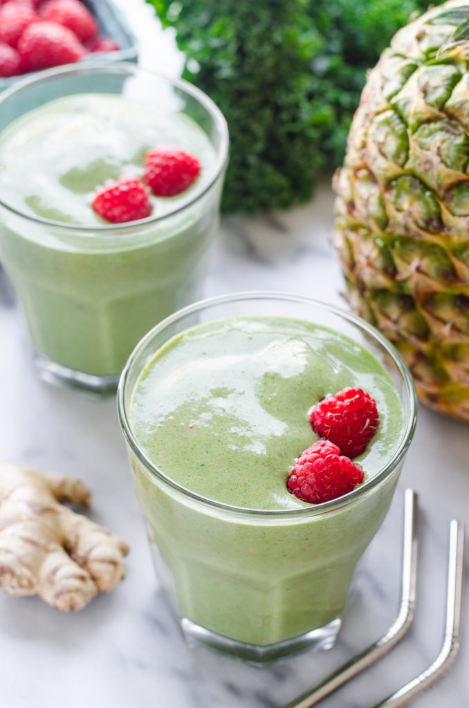 A kale pineapple kefir smoothie with fresh kale and a whole pineapple in the background with fresh ginger and stainless steel straws off to the sides.