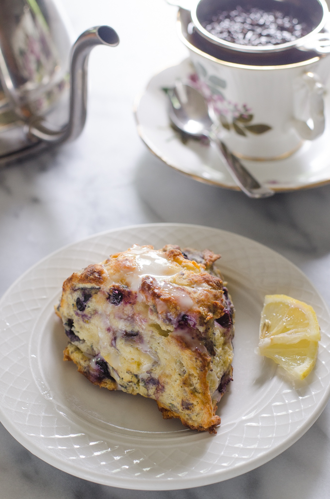 A blueberry Greek yogurt scone on a small plate with mini lemon slices next to it, and a teapot and cup of tea in the background.