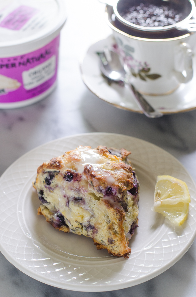 Blueberry scones on a dessert plate with small slices of lemon off to the side and a cup of tea and a tea pot in the background.