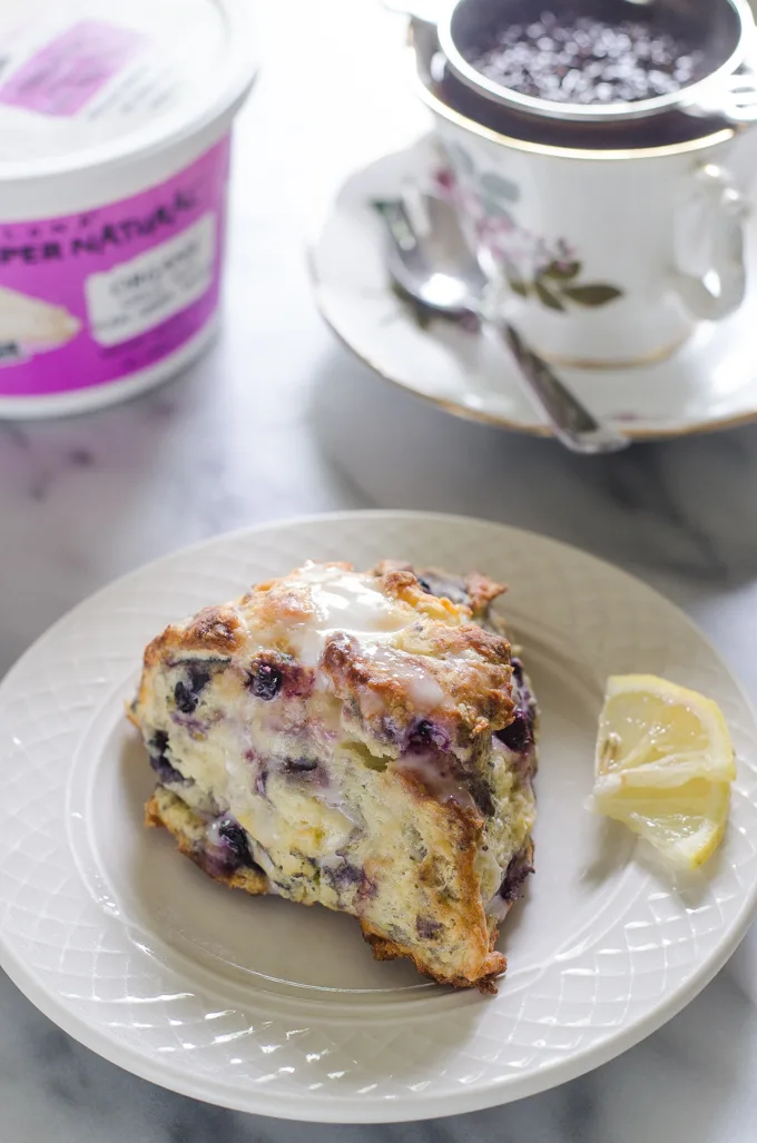 Blueberry scones on a dessert plate with small slices of lemon off to the side and a cup of tea and a tea pot in the background.