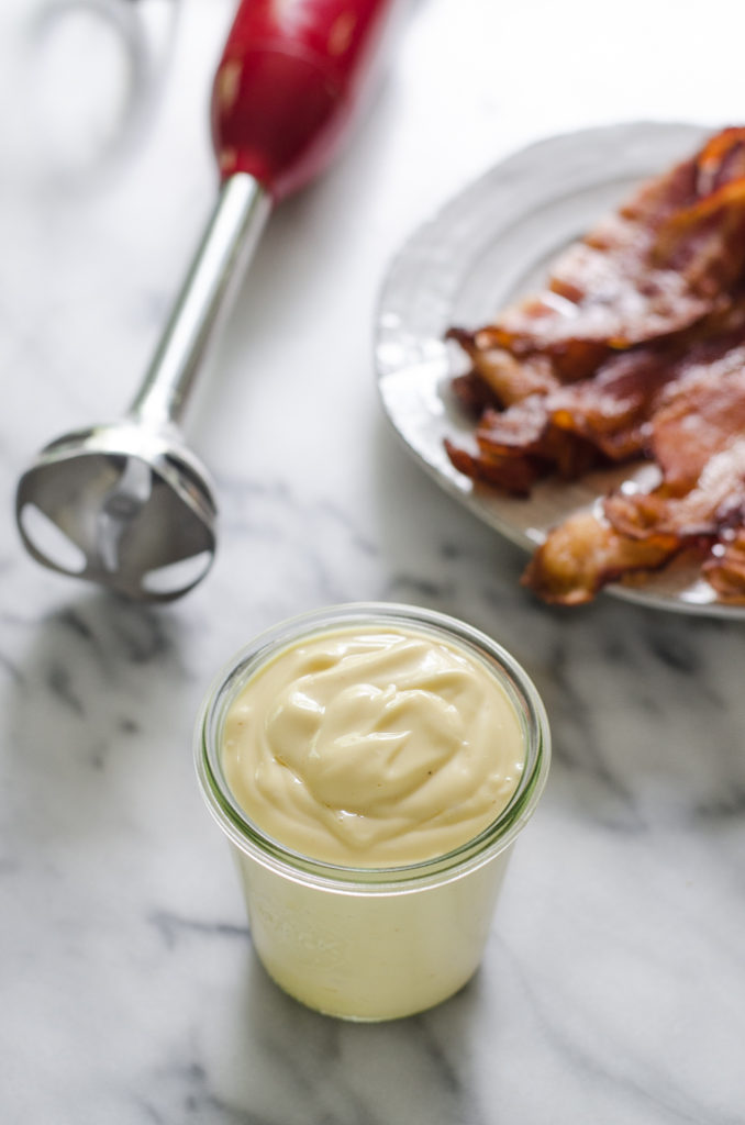 A jar of bacon fat mayo on a marble surface with a plate of cooked bacon and an immersion blender behind it.