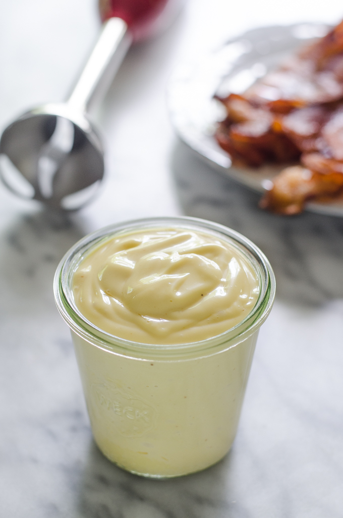 A jar of bacon grease mayonnaise on a marble surface with a plate of cooked bacon behind it.