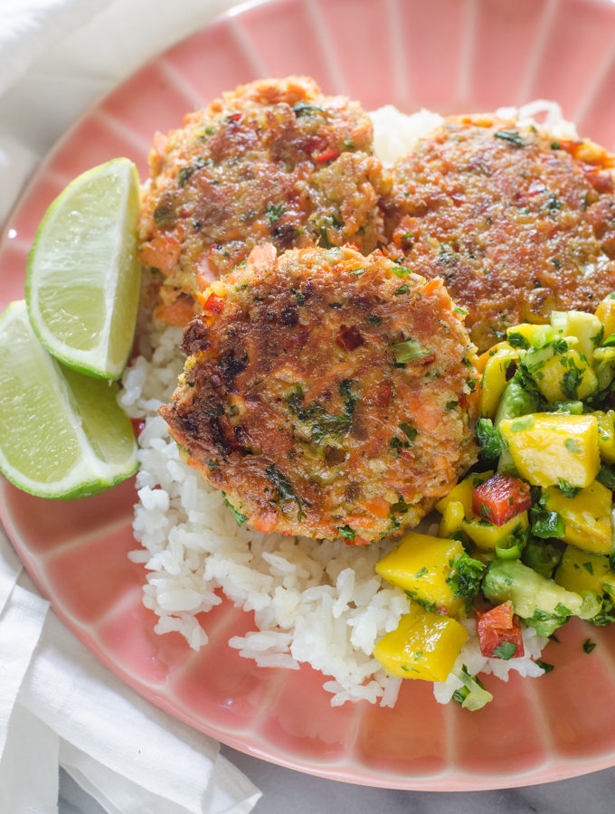 A pink plate with rice, salmon cakes, and mango salsa with lime wedges.