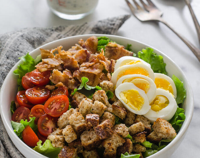 3/4 view shot of BLT salad with dressing and serving utensils in the background on a marble surface.