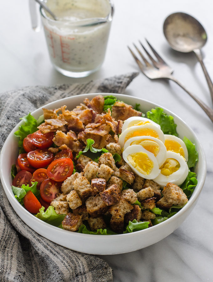 3/4 view shot of BLT salad with dressing and serving utensils in the background on a marble surface.