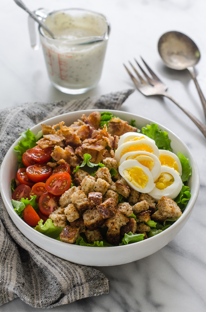 3/4 view shot of BLT salad with dressing and serving utensils in the background on a marble surface.