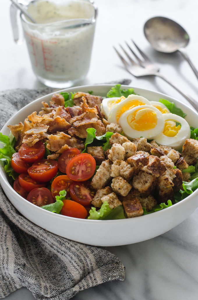 3/4 view shot of BLT salad with dressing and serving utensils in the background on a marble surface.