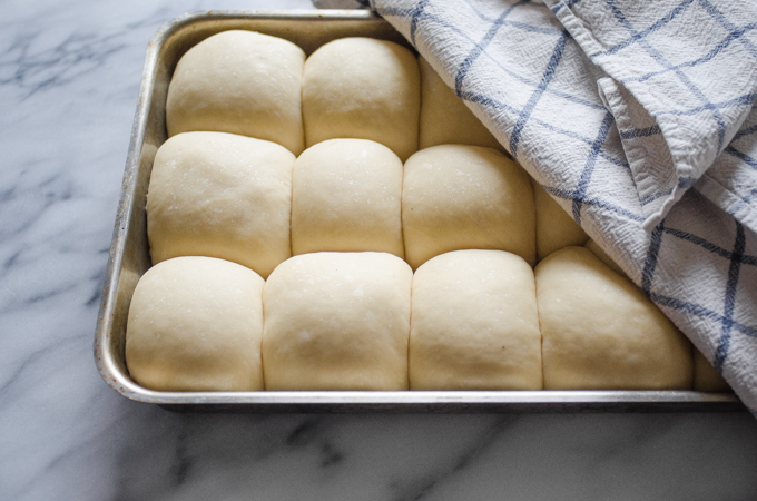 The risen balls of dough in a 13x9 inch stainless steel pan with a checkered tea towel draped over them.