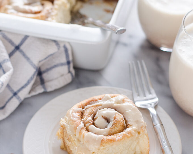 An individual eggnog cinnamon roll on a plate with the pan in the background.