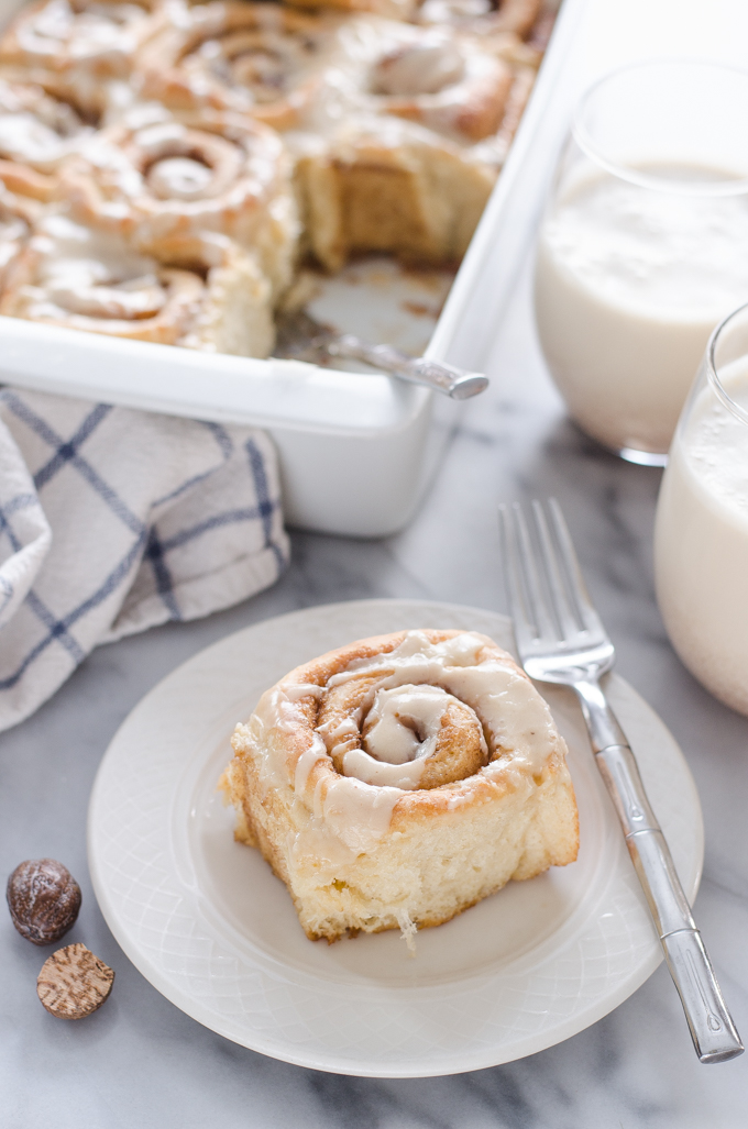An individual eggnog cinnamon roll on a plate with the pan in the background.