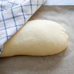 The risen loaf of bread with a tea towel partially covering it.