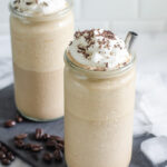 Two cups of mocha frappuccino on a slate board with coffee and ice cubes around it.