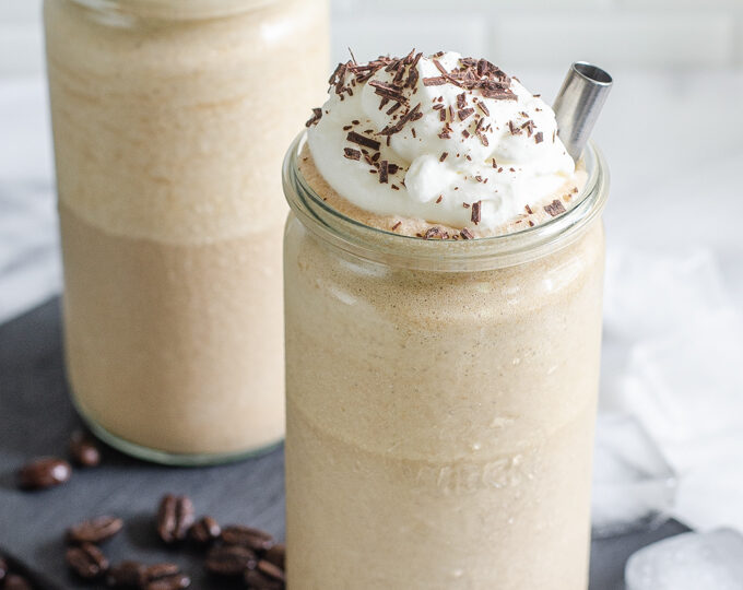 Two cups of mocha frappuccino on a slate board with coffee and ice cubes around it.