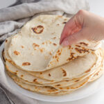 Sourdough tortillas stacked on a plate with a tea towel around them with a hand rolling the top tortilla.