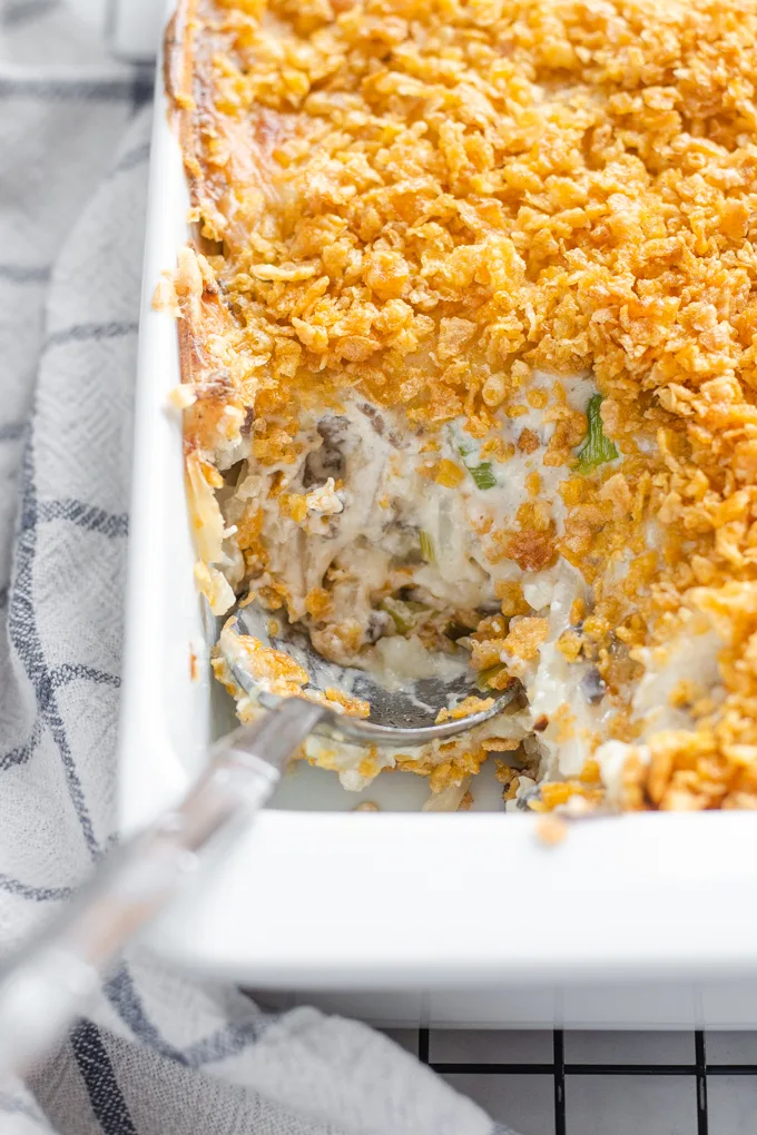 The baked gluten free cheesy potatoes (AKA funeral potatoes) in a white casserole dish on top of a wire cooling rack with a scoop taken out.