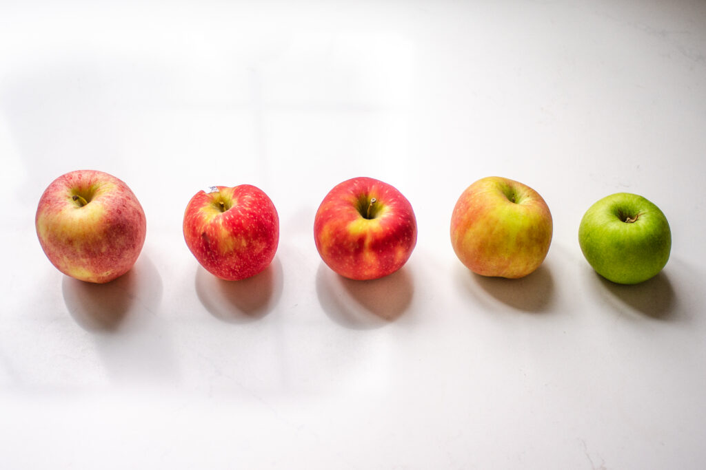 Different kinds of apples lined up on a counter: Gala, Pink Landy, Honeycrisp, Fuji, and Gala.