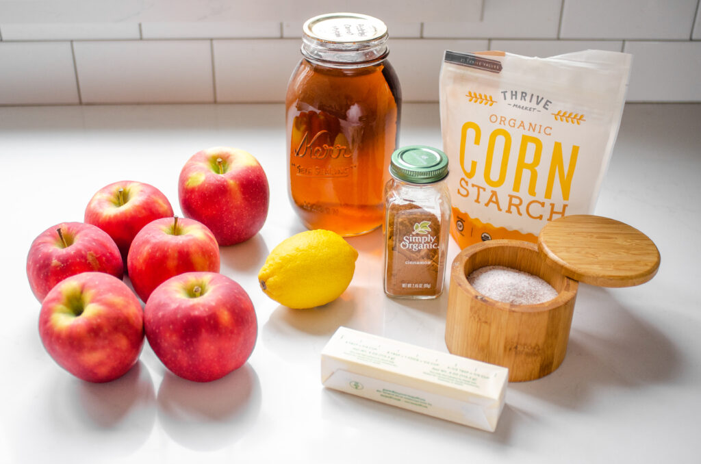 All of the ingredients need to make baked cinnamon apples laid out on a counter: apples, lemon, maple syrup, cornstarch, salt, butter, and cinnamon. 