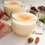 Two mugs of homemade eggnog with a tea towel, greenery, and fresh nutmeg around them.