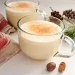 Two mugs of homemade eggnog with a tea towel, greenery, and fresh nutmeg around them.
