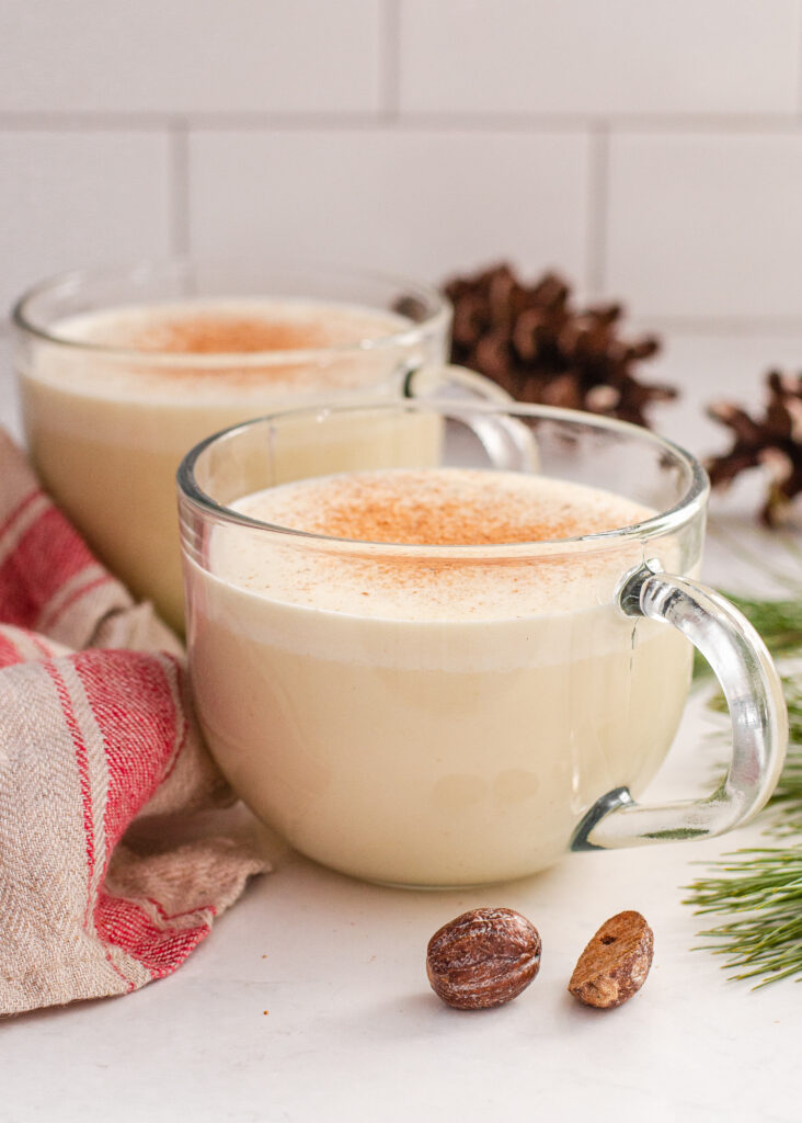 Two mugs of my homemade cooked eggnog recipe without alcohol with a tea towel, greenery, and fresh nutmeg around them.