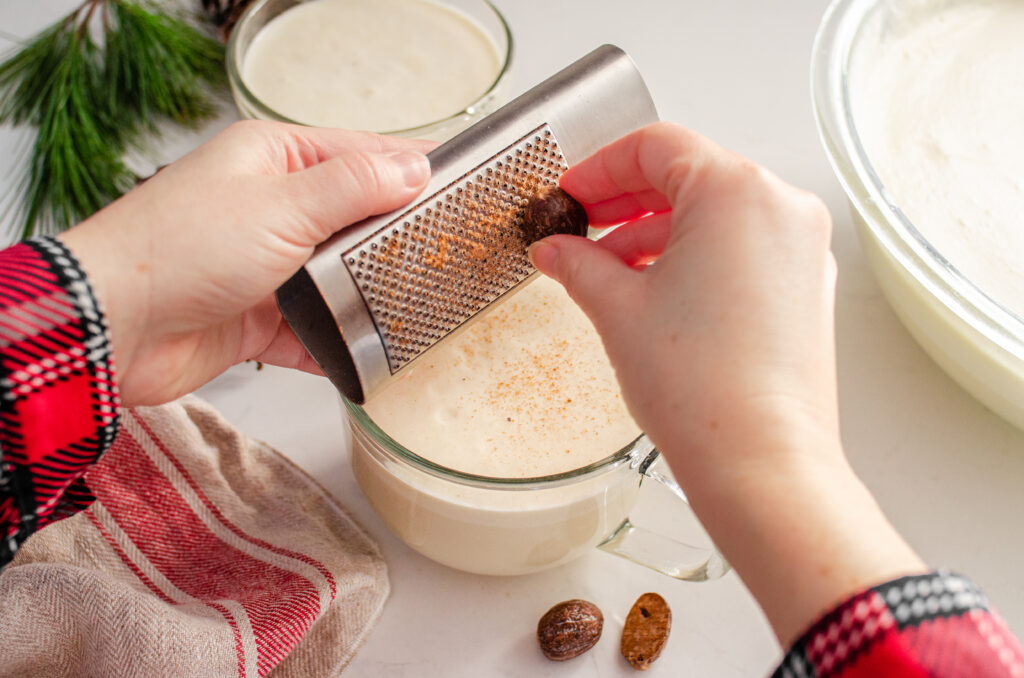Grating fresh nutmeg over the mugs of my homemade cooked eggnog recipe without alcohol.
