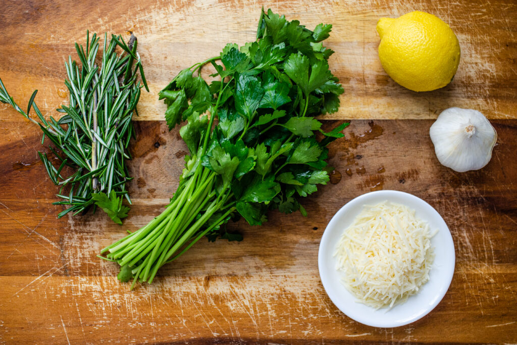Fresh rosemary, fresh parsley, a lemon, a garlic bulb, and parmesan cheese on a cutting board.