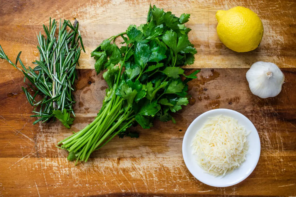 Fresh rosemary, fresh parsley, a lemon, a garlic bulb, and parmesan cheese on a cutting board.